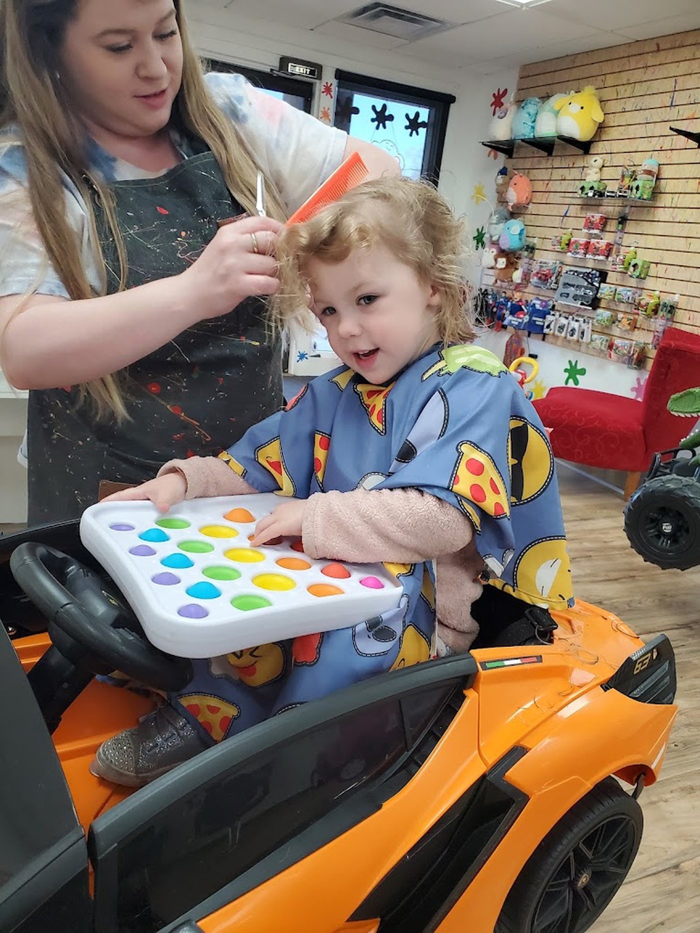 A toddler in an orange ride-on car holding a pop-it tray while a stylist trims her hair.