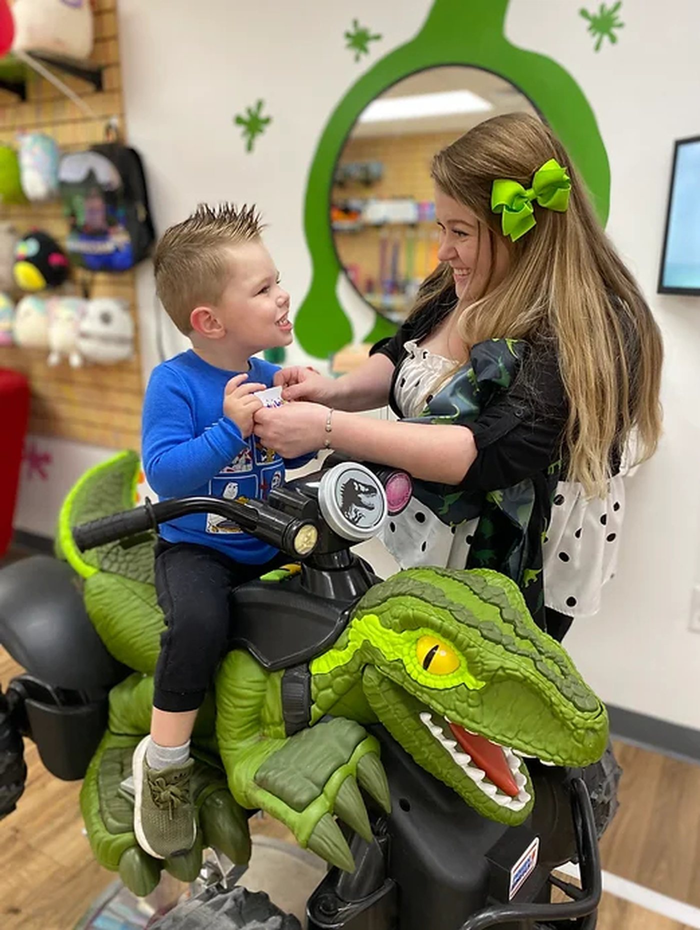 A boy sitting on a large green ride-on dinosaur, laughing with his stylist, in front of a wall painted with a green paint splat.